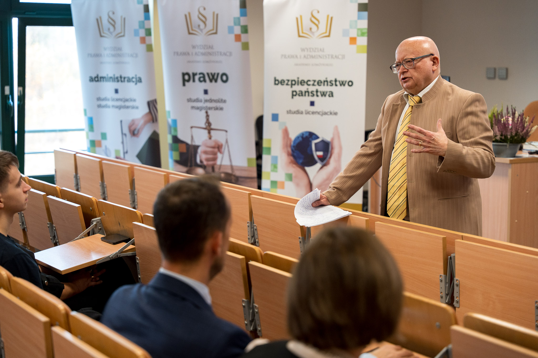 Mężczyzna w garniturze w paski i żółtym krawacie prowadzi wykład w sali uniwersyteckiej. Stoi przy pierwszym rzędzie ławek, trzymając kartki z notatkami. Przed nim siedzi kilku słuchaczy. W tle widoczne są roll-upy z napisami po polsku dotyczącymi kierunków studiów: administracja, prawo oraz bezpieczeństwo państwa.