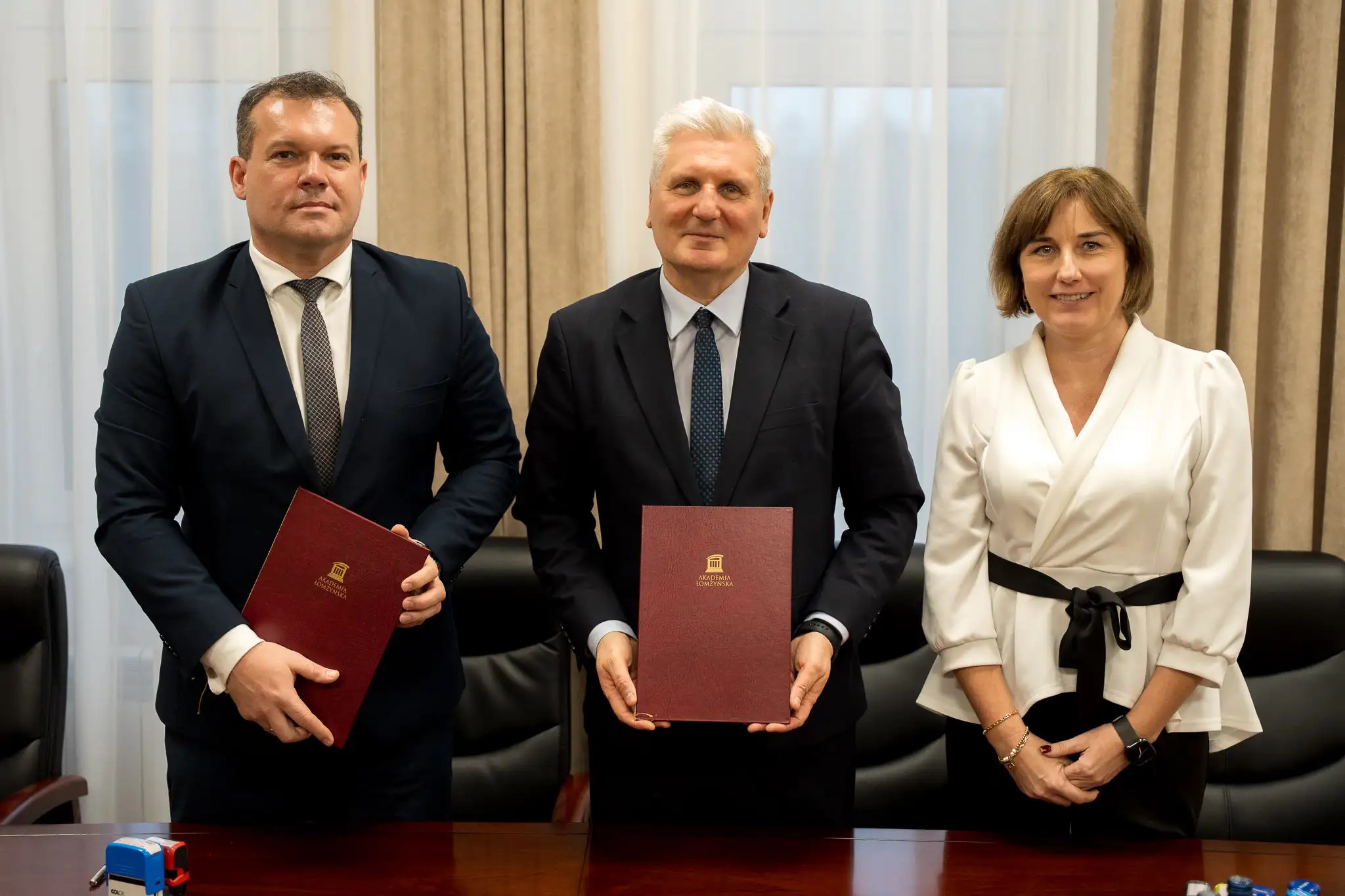 Kobieta i dwaj mężczyźni stoją za stołem konferencyjnym. Dwaj mężczyźni trzymają w ręku teczki z dokumentami.