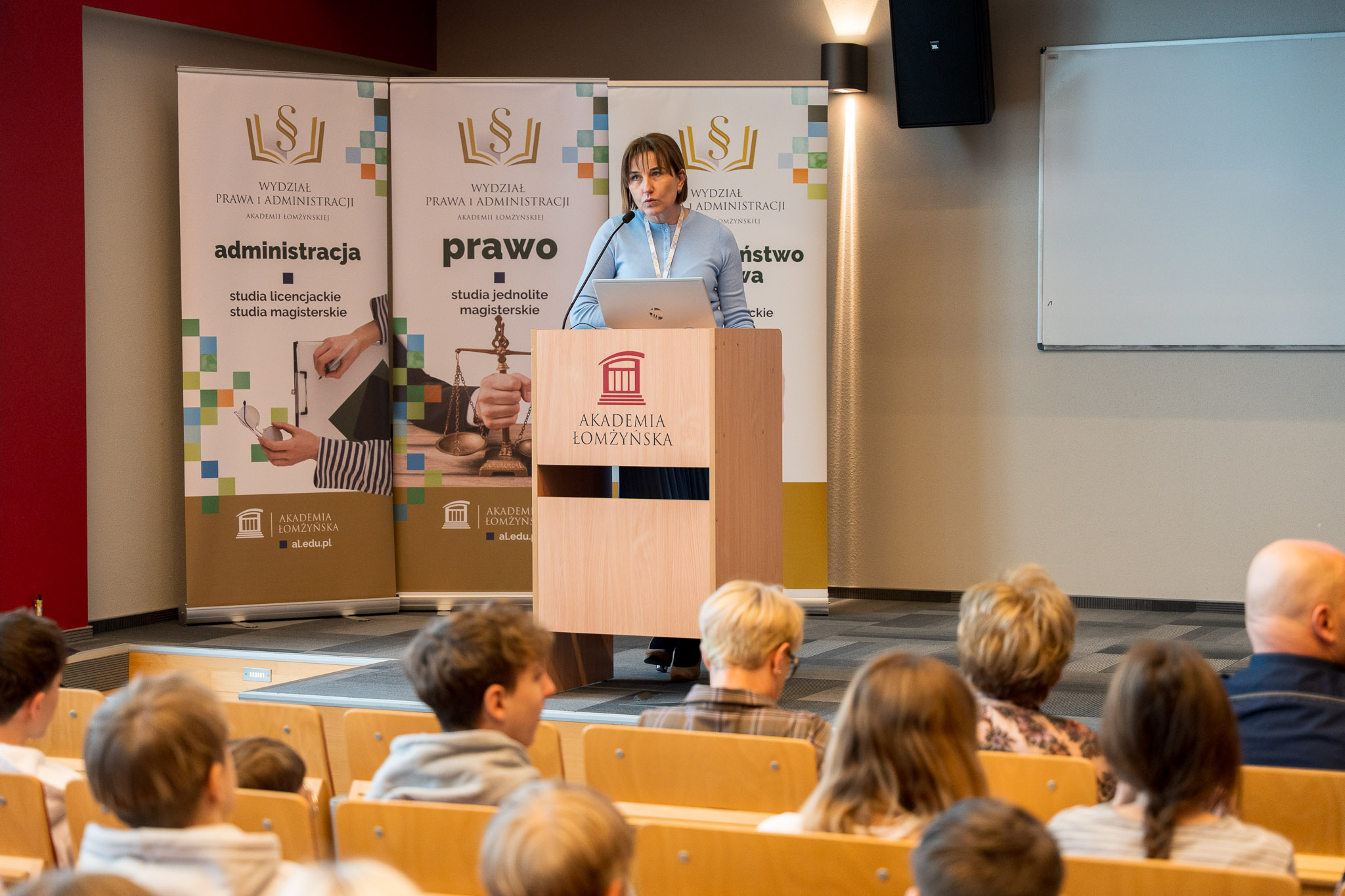 Kobieta stojąca za mównicą przemawia do osób siedzących na krzesłach w auli wykładowej.