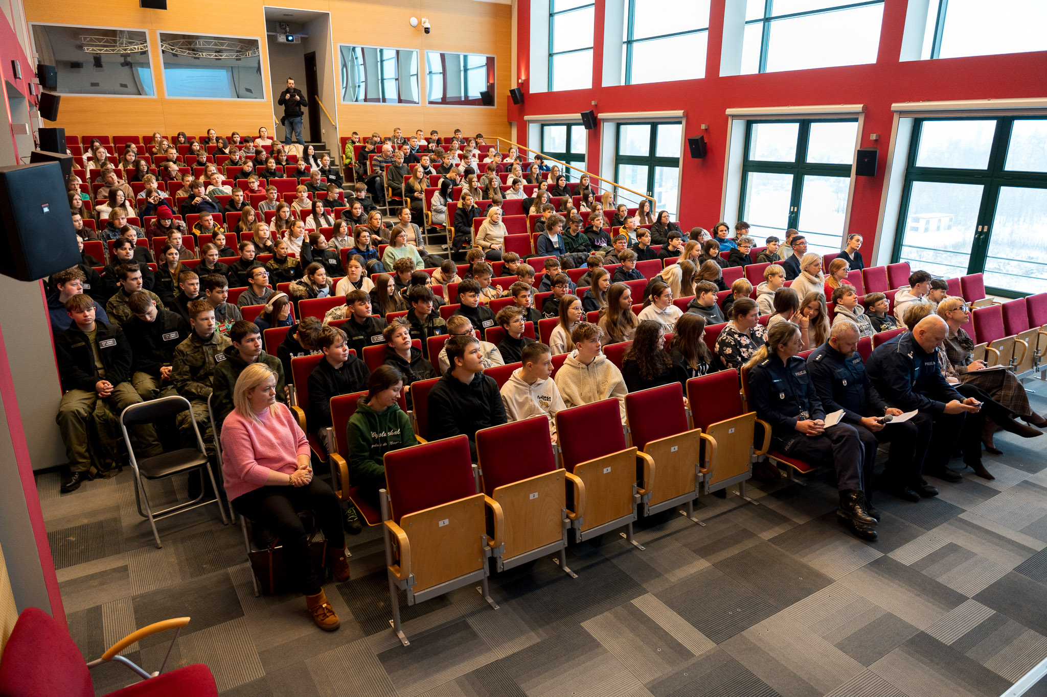 Osoby siedzące na krzesłach w auli wykładowej, słuchają uważnie.