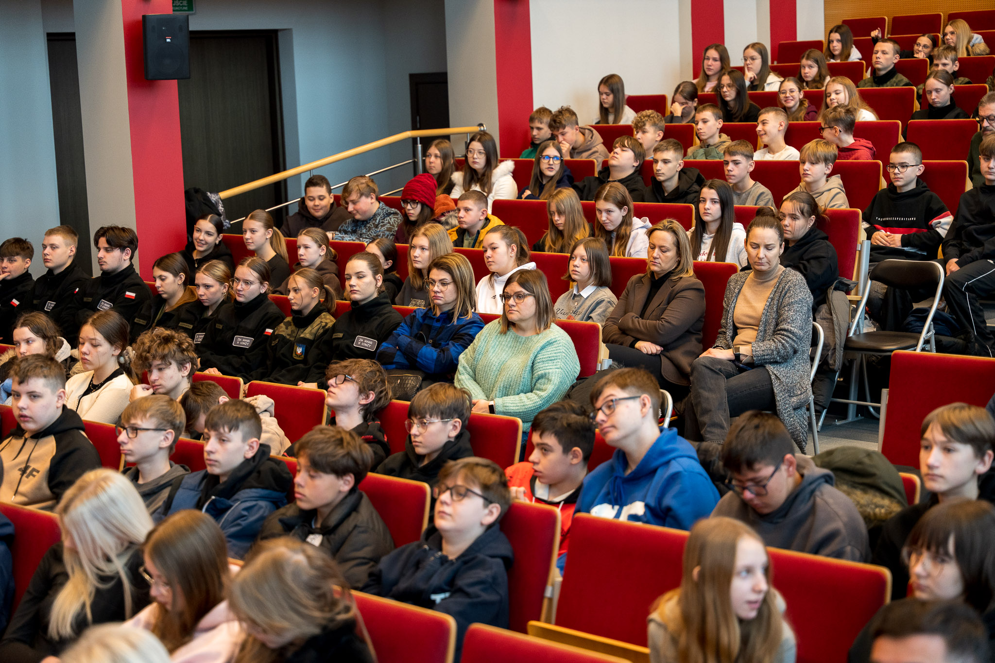 Grupa osób słucha uważnie siedząc na krzesłach w auli wykładowej.