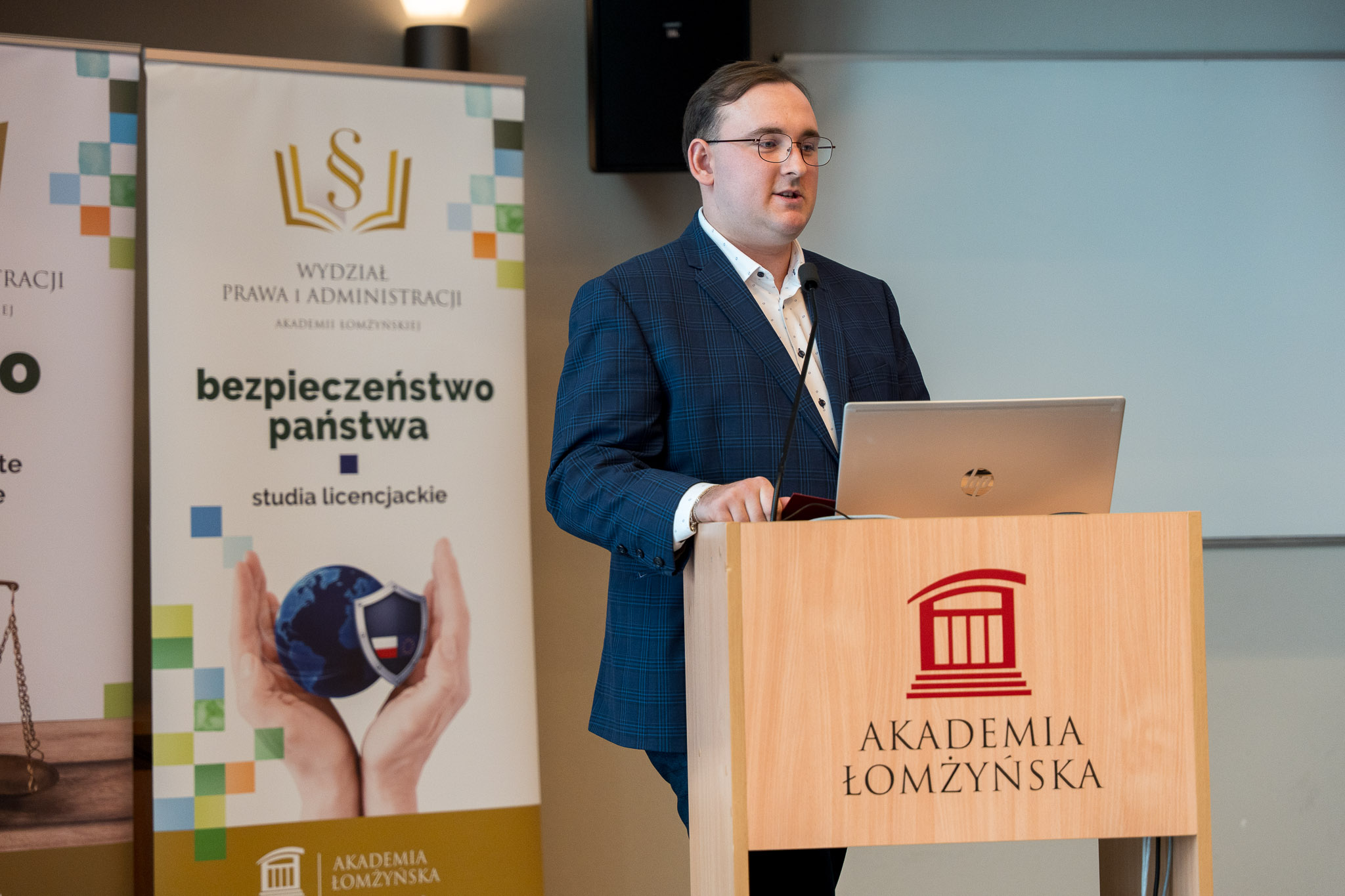 Mężczyzna stojący za mównicą przemawia na podeście auli wykładowej, w tle roll-up kierunku bezpieczeństwa państwa.