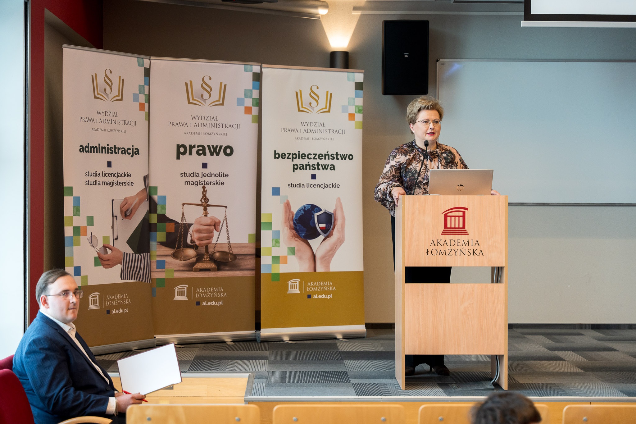 Kobieta stojąca za mównicą przemawia na podeście auli wykładowej. Z boku na krześle siedzi mężczyna.
