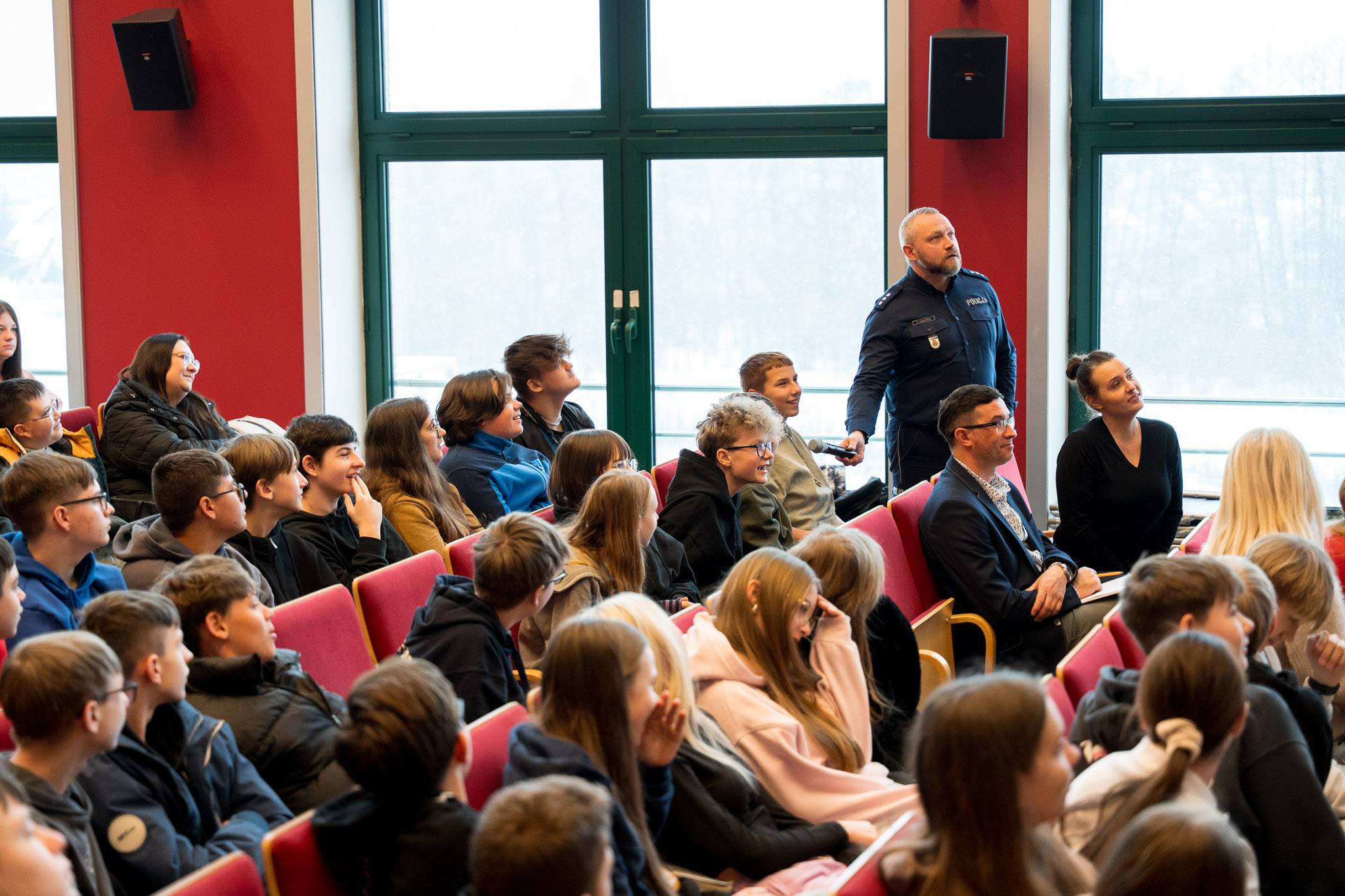 Osoby siedzące na krzesłach w auli wykładowej patrzą na stojącego mężczyznę w mundurze policjanta.