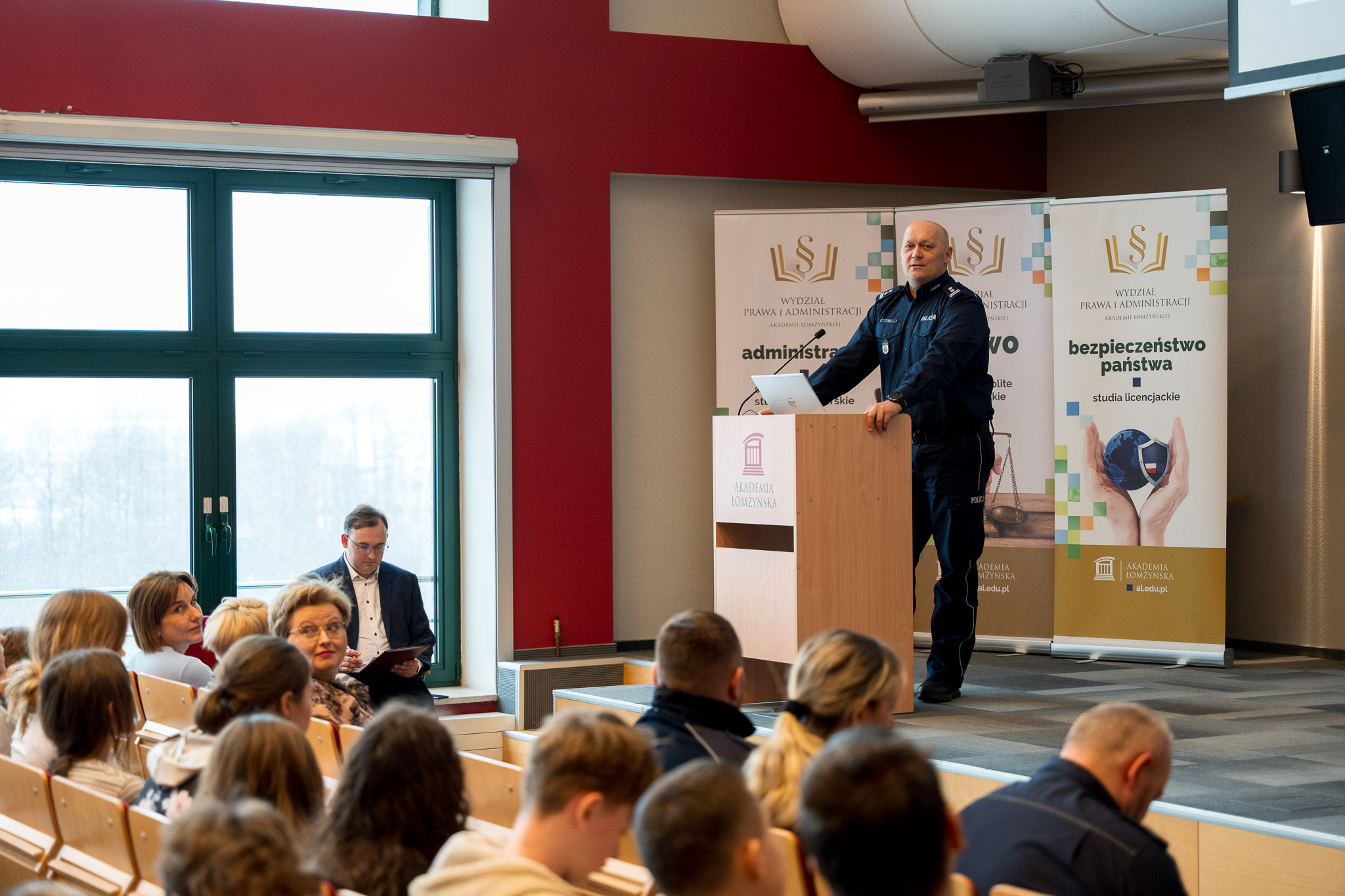 Mężczyzna w mundurze policjanta stojący za mównicą przemawia na podeście auli wykładowej.