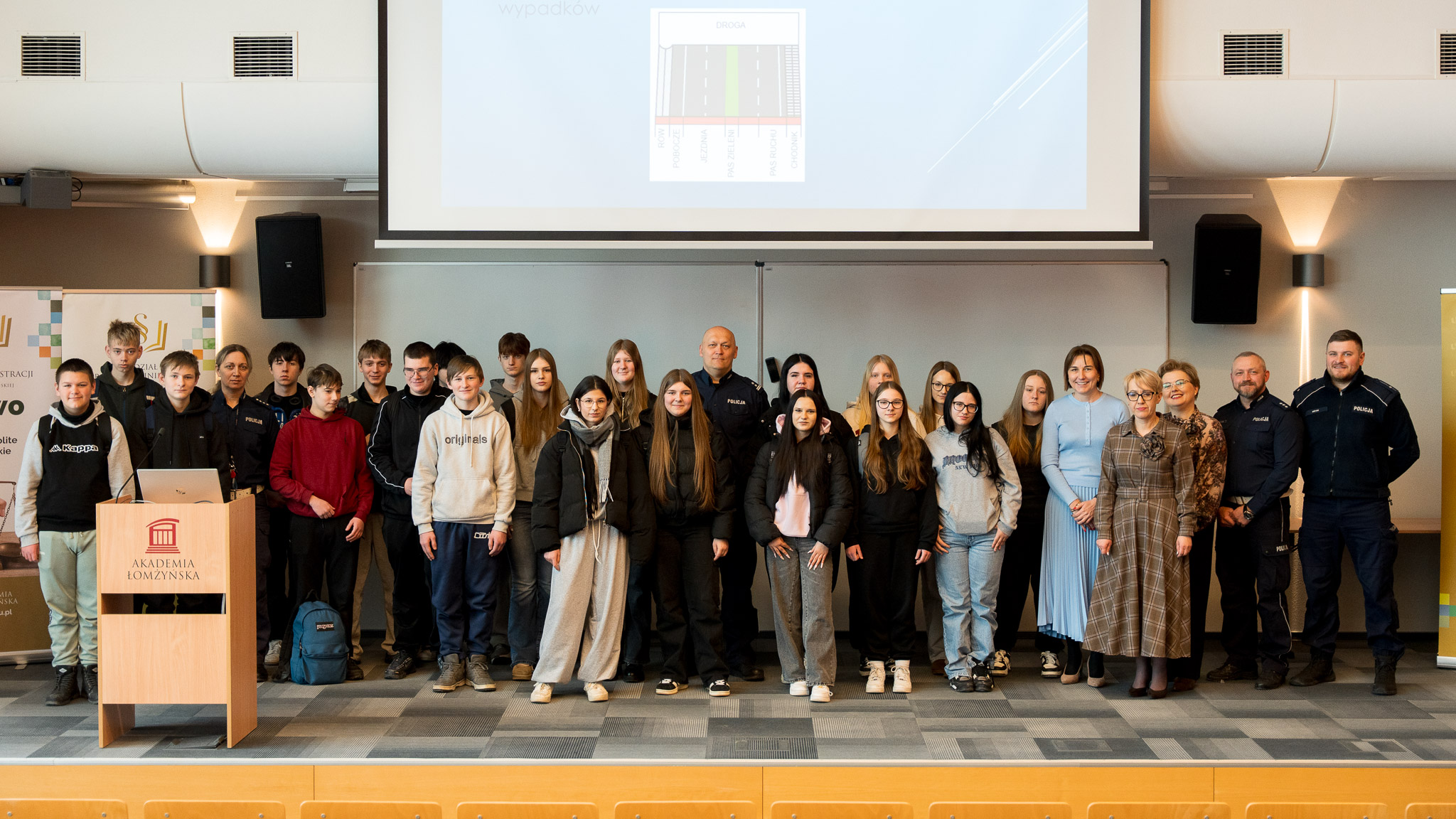 Grupa osób pozuje do zdjęcia na podeście auli wykładownej, w tle ekran projekcyjny.