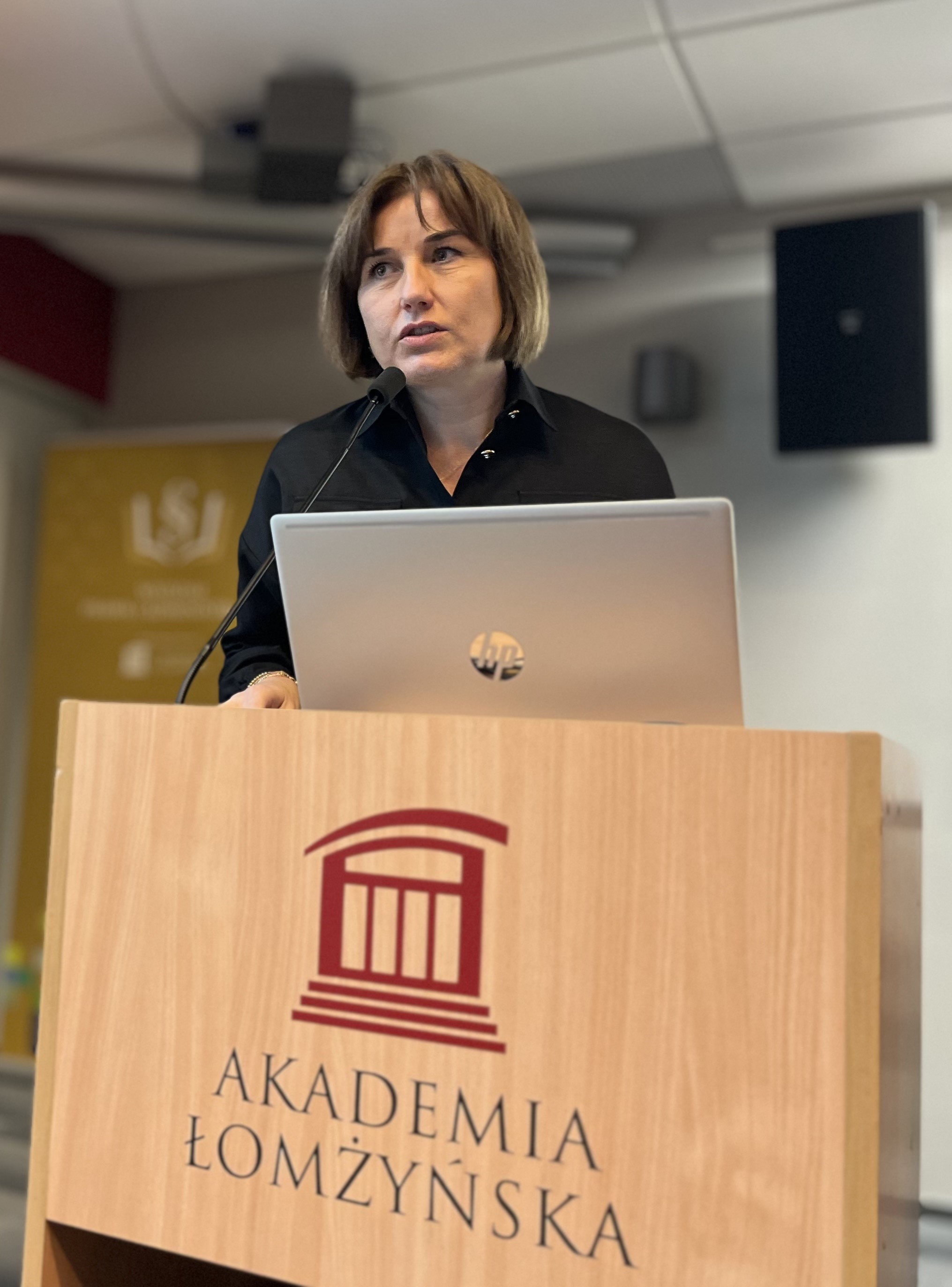 Kobieta stoi za mównicą z napisem „Akademia Łomżyńska” i prowadzi prezentację przy użyciu laptopa. W tle widoczny jest baner uczelni.