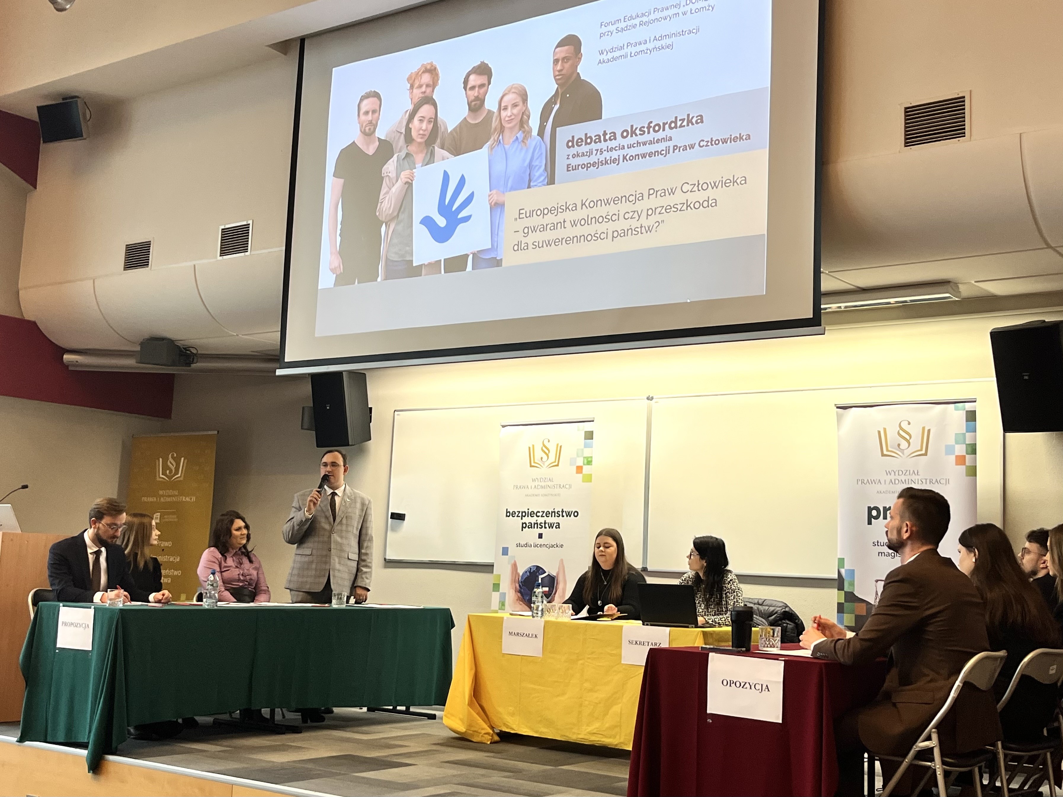 Grupa osób siedzi za stołami nakrytymi obrusami. Jeden mężczyzna stoi przemawiając przez mikrofon.