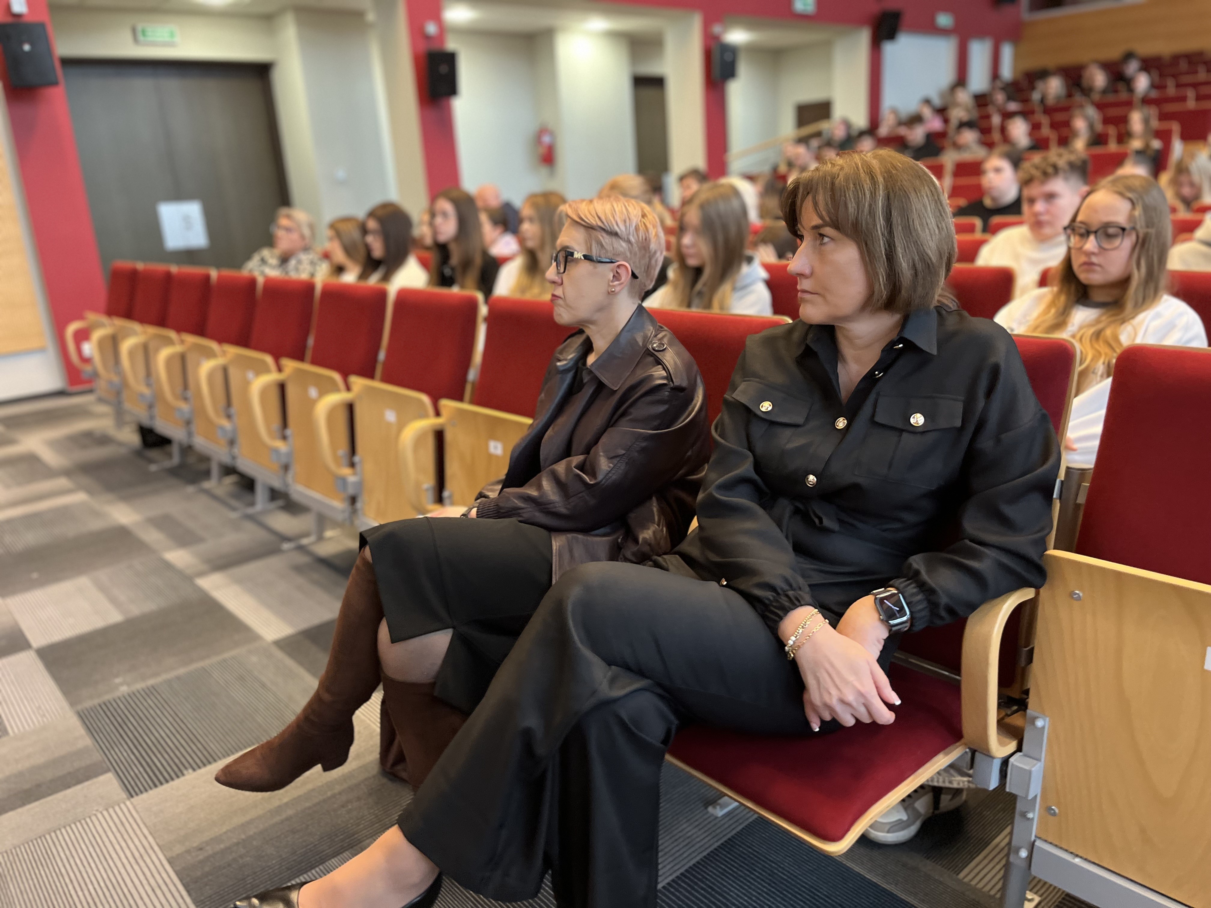 Osoby siedzą w sali wykładowej. W pierwszym rzędzie widać dwie kobiety, a za nimi znajduje się kilka rzędów wypełnionych uczestnikami.