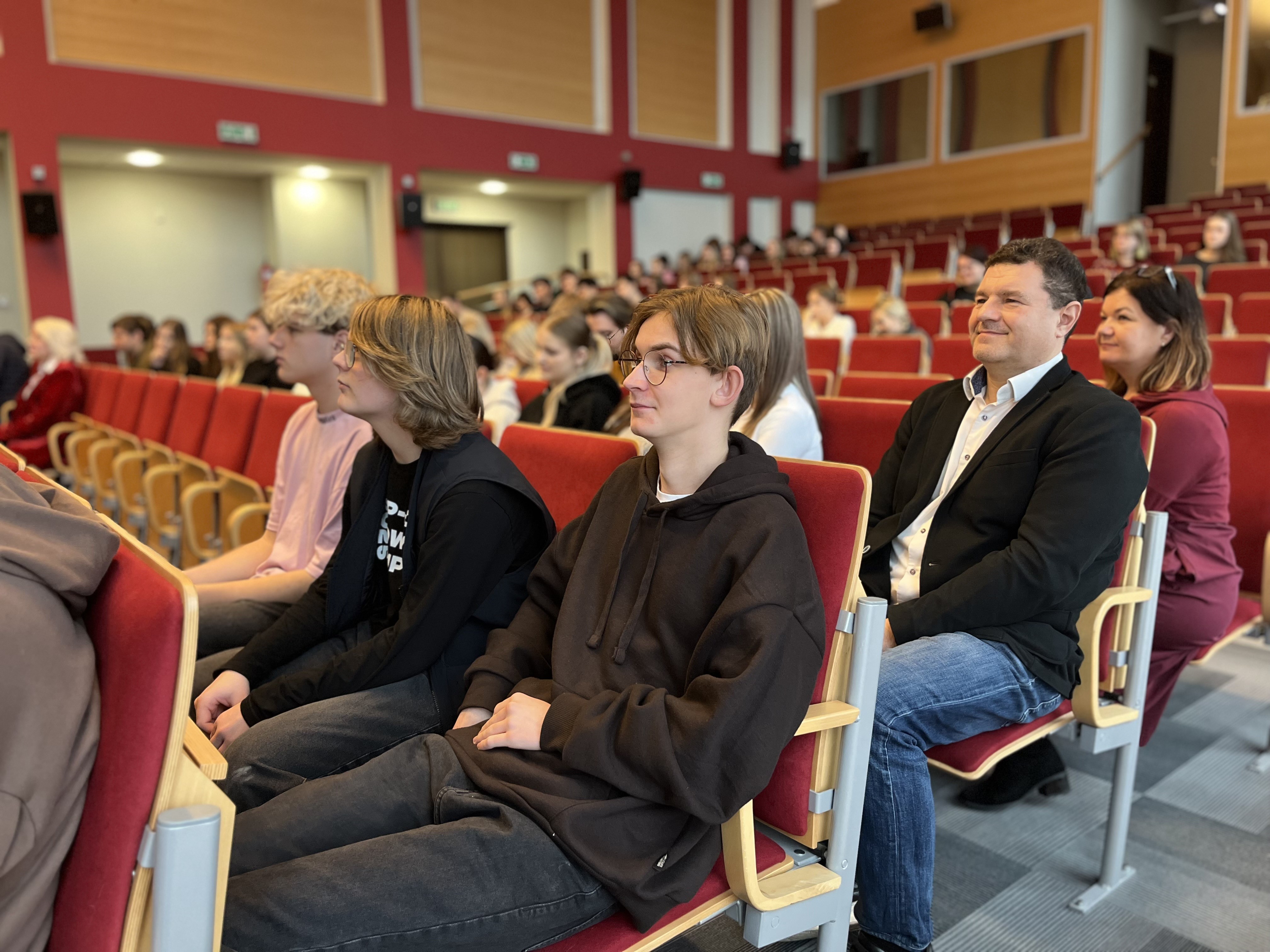 Uczestnicy spotkania siedzą w auli wykładowej uważnie słuchając.