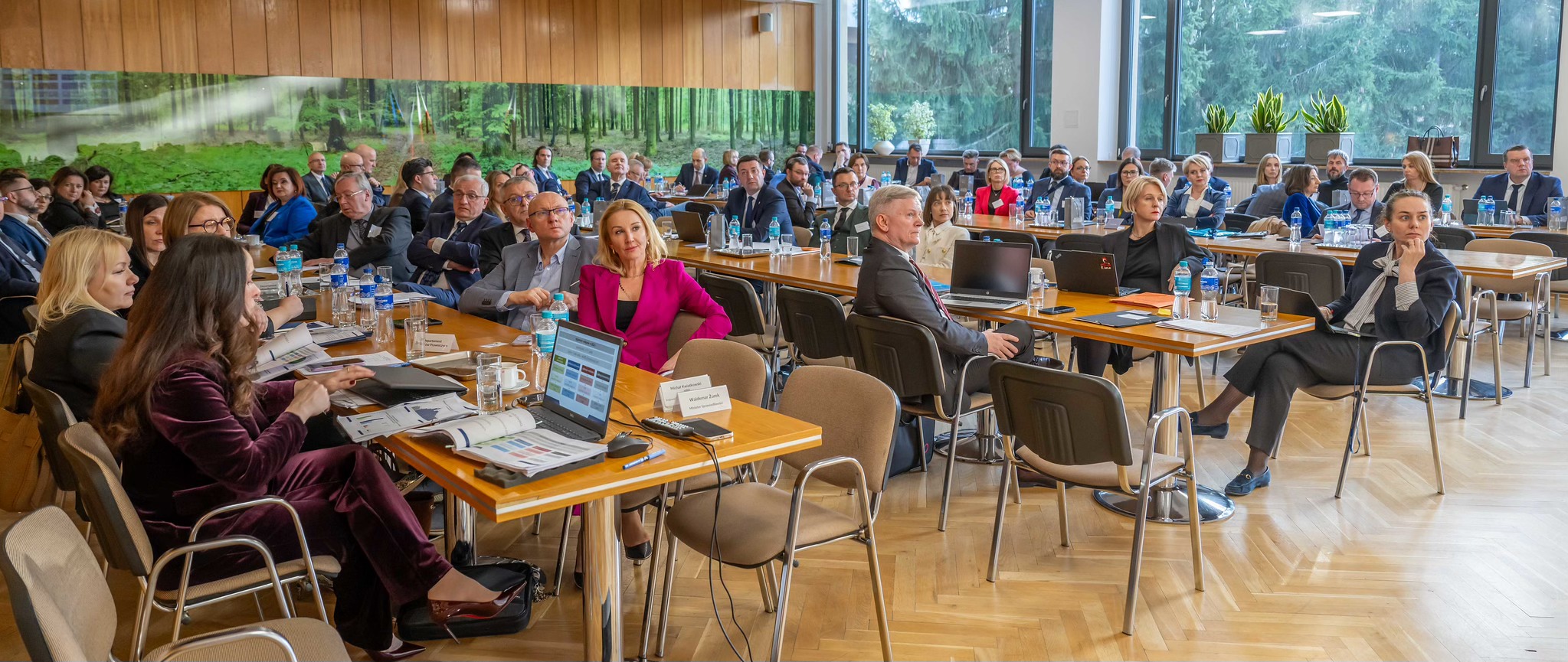 Grupa ludzi siedzi na krzesłach za stołami konferencyjnymi