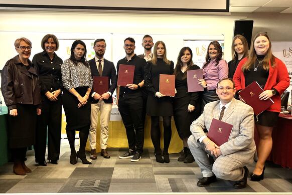 Grupa osób pozuje do zdjęcia podczas wydarzenia akademickiego. Część trzyma teczki na dokumenty z emblematem uczelni.